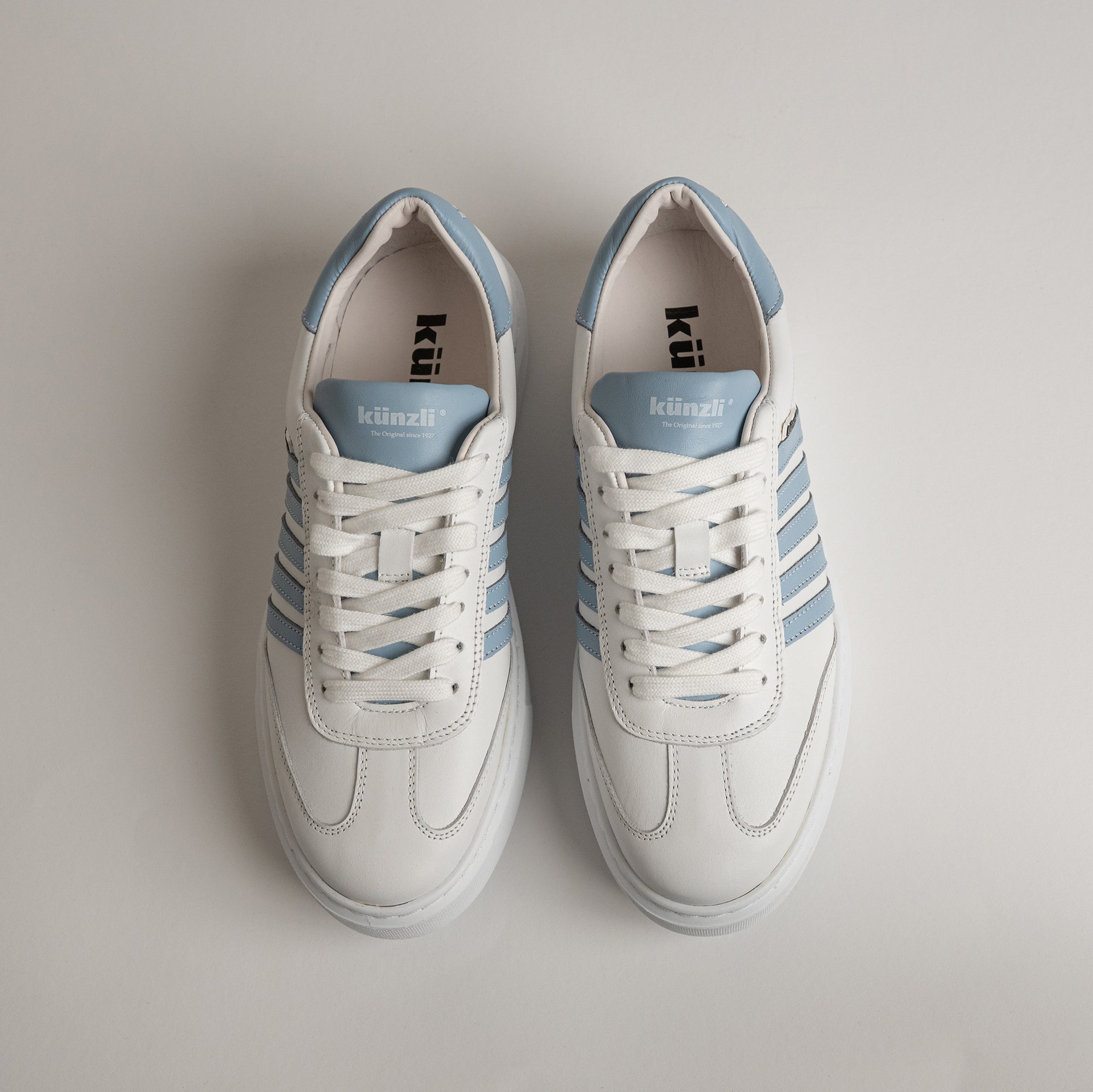 Weiße Kuenzli Schuhe mit blauen Streifen auf hellem Hintergrund, sportlich und modern.