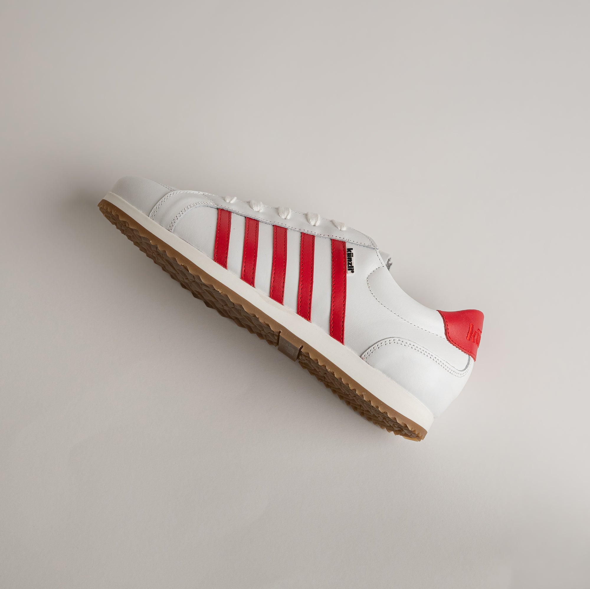 Weißer Kuenzli Schuh mit roten Streifen und Gummisohle auf grauem Hintergrund.